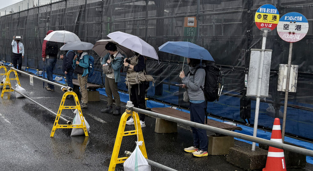 雨のなか傘を手に路線バスの到着をじっと待つ観光客ら＝2025年11月5日、屋久島空港、屋久島ポスト撮影