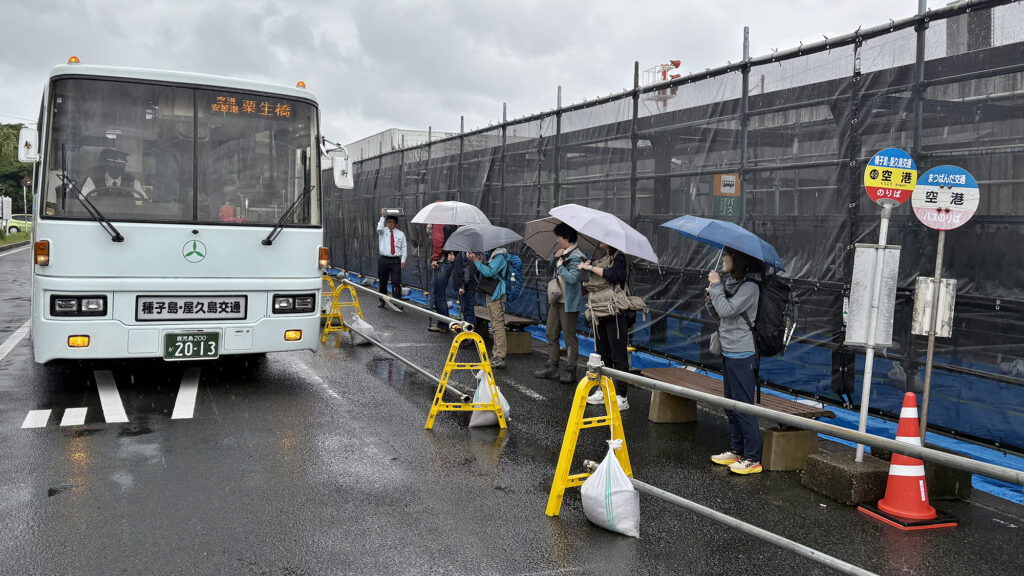 雨に打たれながら傘を手に路線バスを待つ観光客ら＝2025年11月5日、屋久島空港、屋久島ポスト撮影